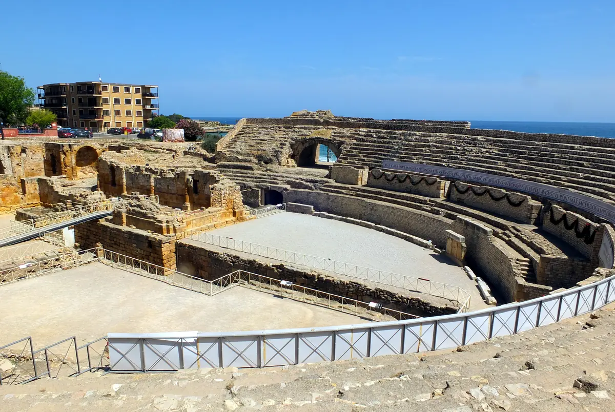 The ruins of a Roman amphitheatre seen from above, with stone seating rising in tiers and the sea beyond