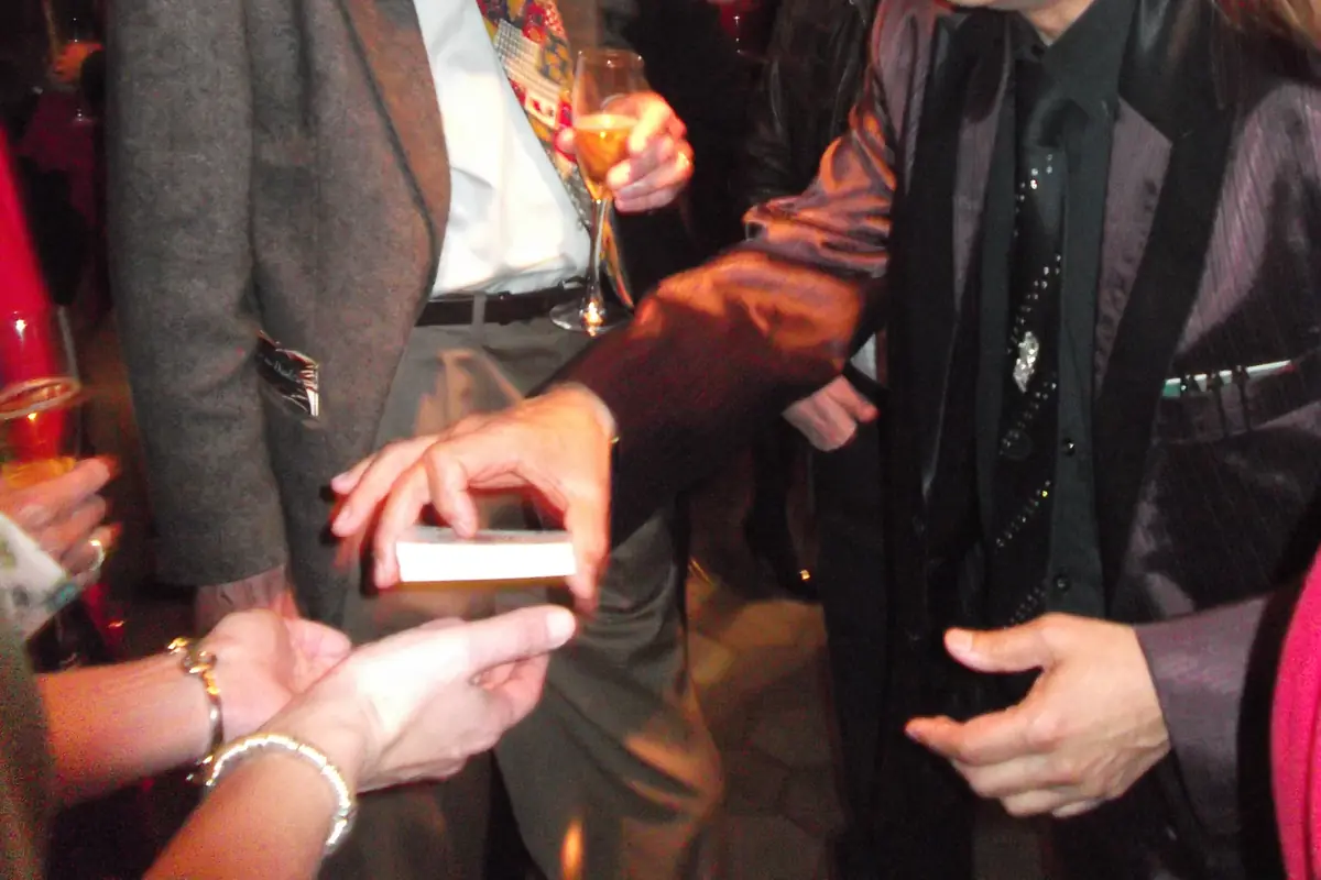 A magician's hands holding a small stack of playing cards at a restaurant table, with a guest's hands reaching in from the lower left