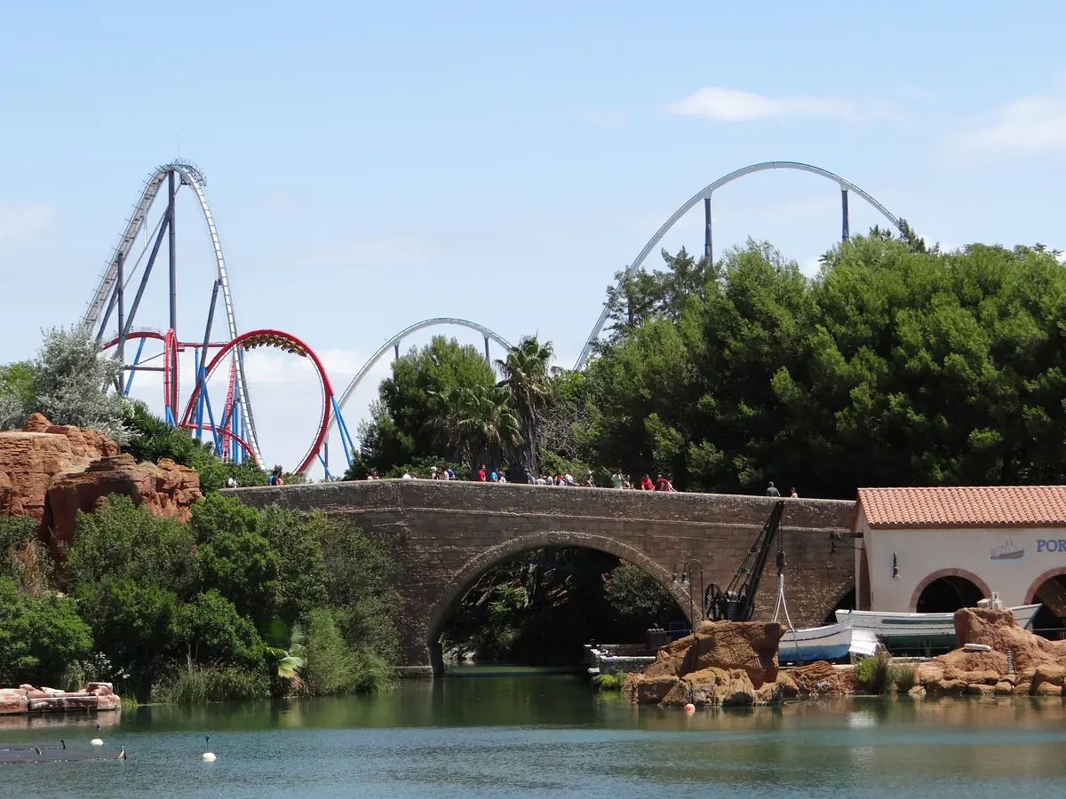 A stone bridge crosses a pond beside red rock formations, with roller coaster tracks rising behind trees