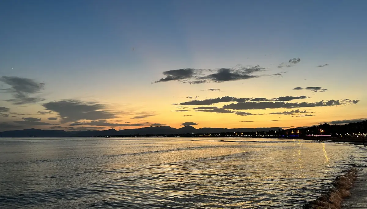 A calm sea reflects a yellow and orange sunset sky, with the lit shoreline of Salou along the right edge