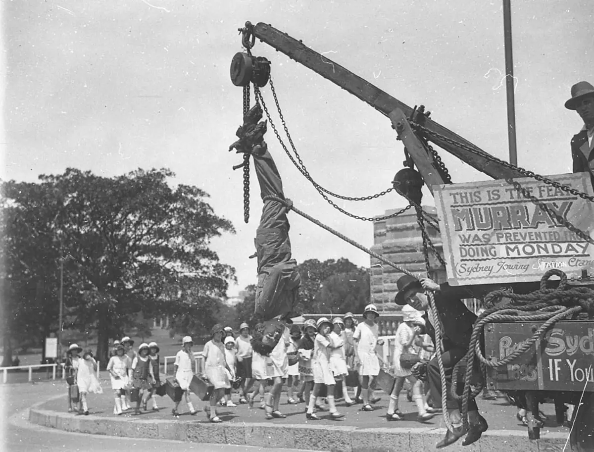 A man hangs upside down in a straitjacket from a crane in a street, with a large crowd and a sign below him