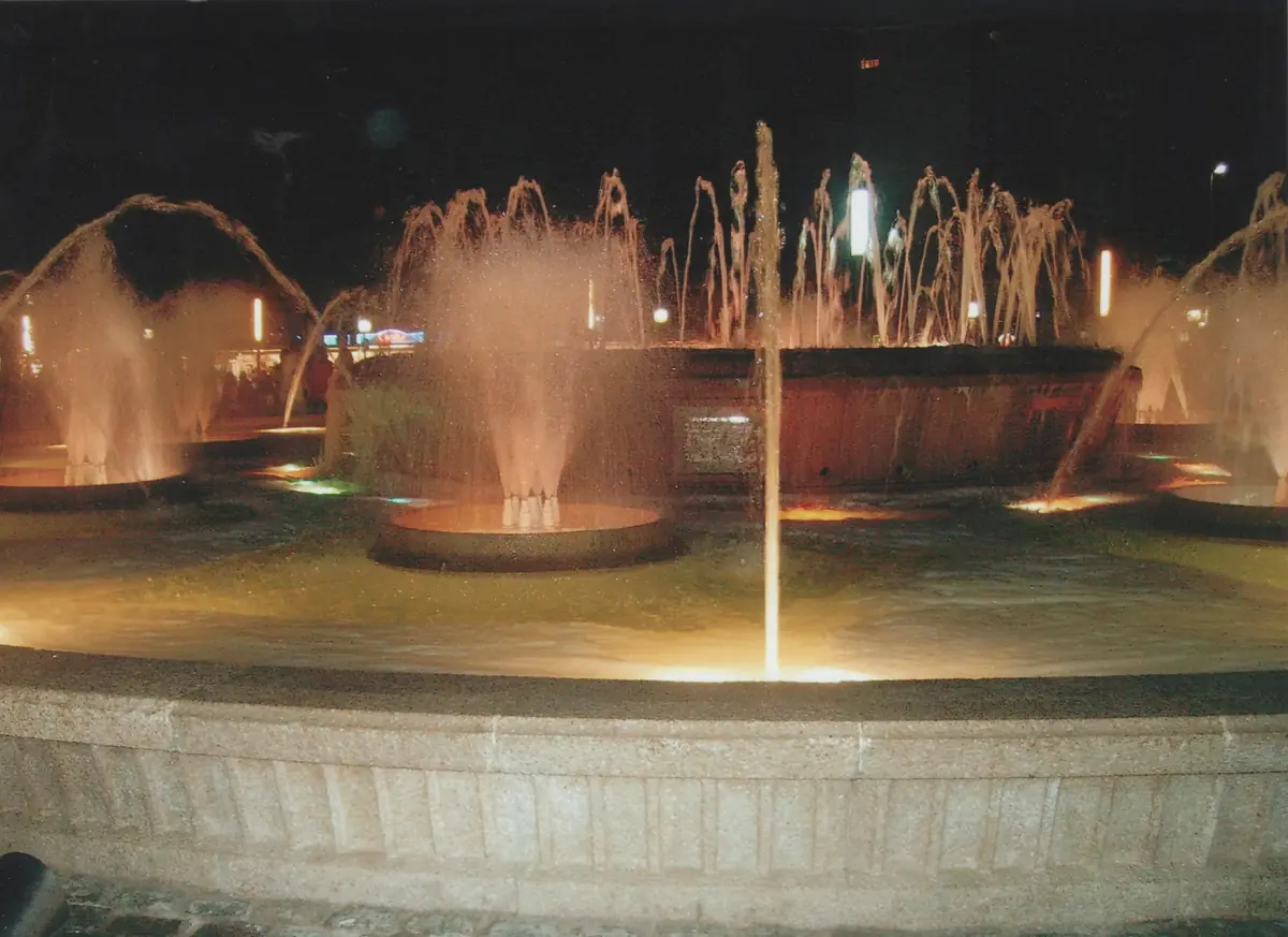 Several jets of water rise from a round fountain lit orange against a dark night sky, with a low stone rim in the foreground