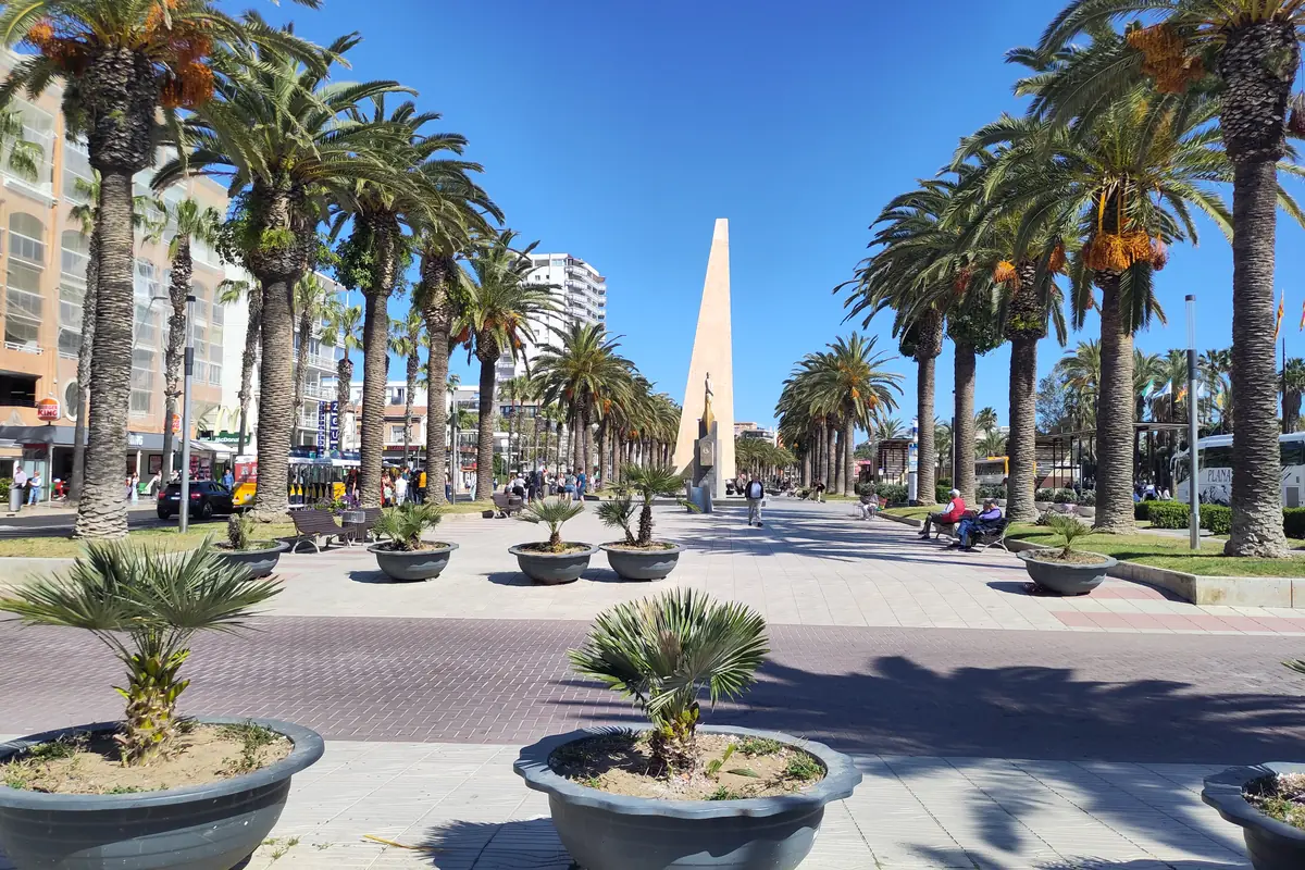 A wide paved promenade lined with tall palm trees and planters leads towards a tall slim obelisk under a blue sky
