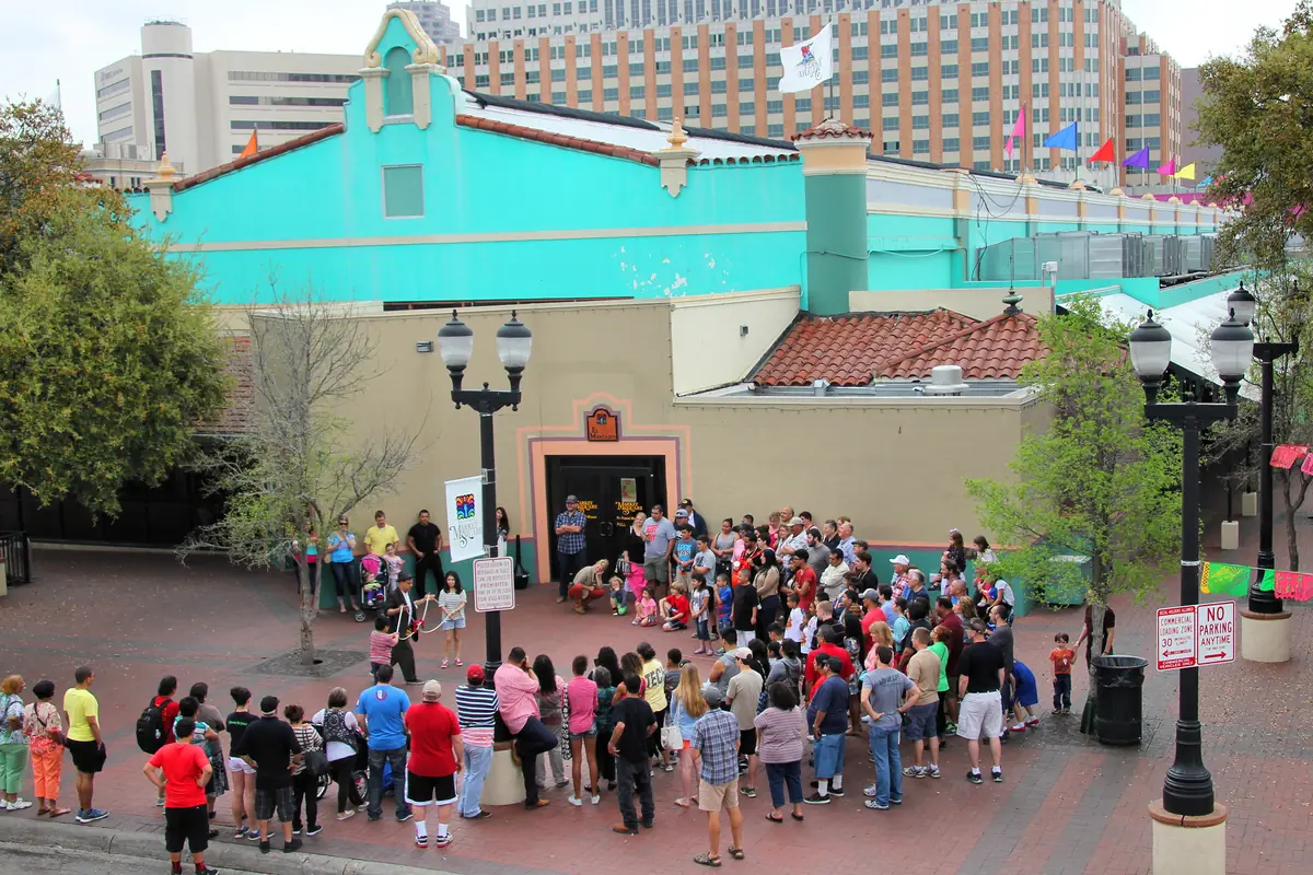 A magician performing for a large crowd standing in a tiled market square in front of a turquoise building, seen from above