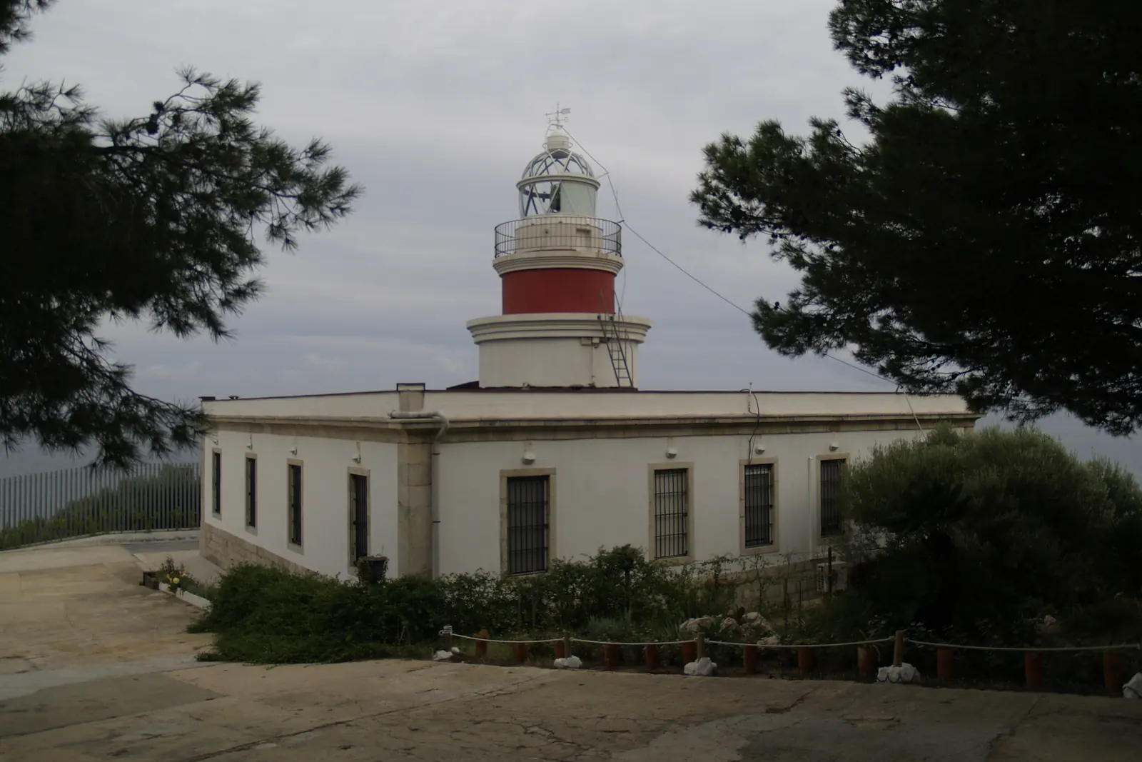 The Salou lighthouse, a white building with a red and white tower, set among pine trees under a cloudy sky