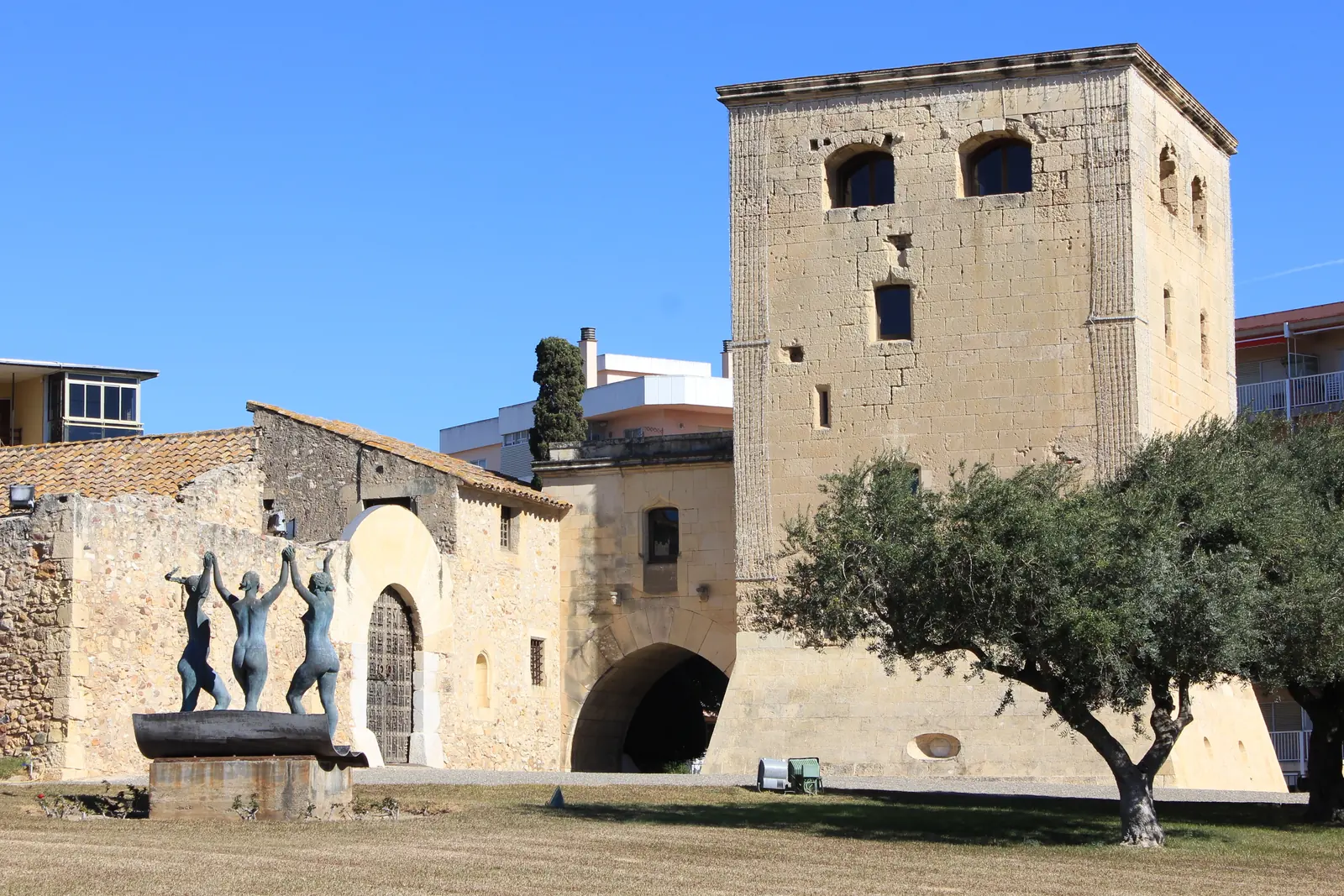 The Torre Vella in Salou, a square stone tower with a stone building beside it and a sculpture of three figures in front under a blue sky